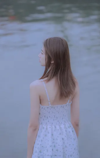 一位穿着白色连衣裙的年轻女子沿着海滩走向水边 注视着水面。