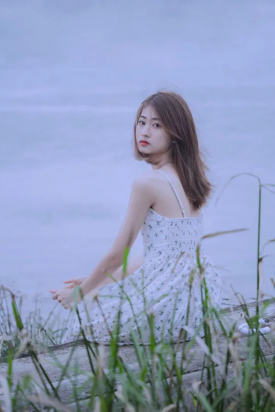 一位穿着白色连衣裙的年轻女子坐在湖边草地上