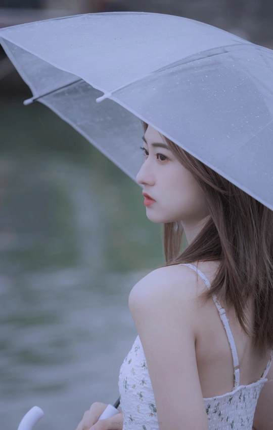 一位年轻女子手持透明雨伞站在雨中的水边