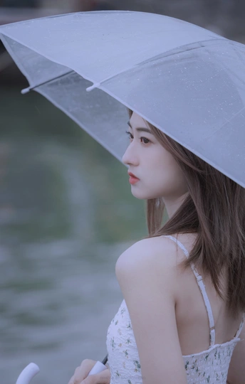 一位年轻女子手持透明雨伞站在雨中的水边