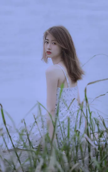 一个穿着白裙子的年轻女子坐在水边草地上