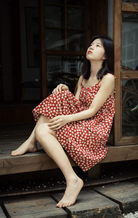 一个穿着红裙子的年轻女子坐在木质台阶上 位于窗户前。