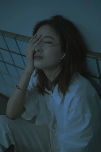 一个年轻女子穿着白色衬衫坐在床上 闭着眼睛 双手放在头上。