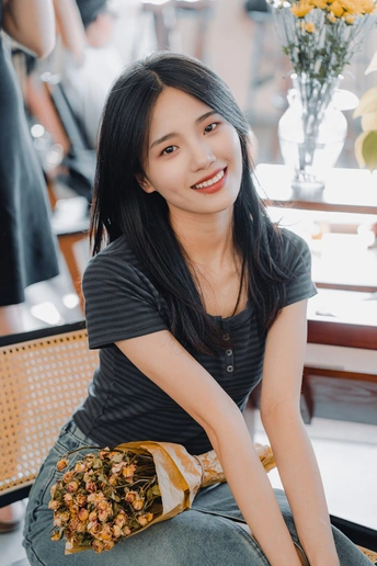 一位年轻女子抱着花坐在桌子旁的椅子上 对着相机微笑。
