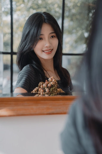 一位年轻女子坐在桌子前 前方有花 正在照镜子。