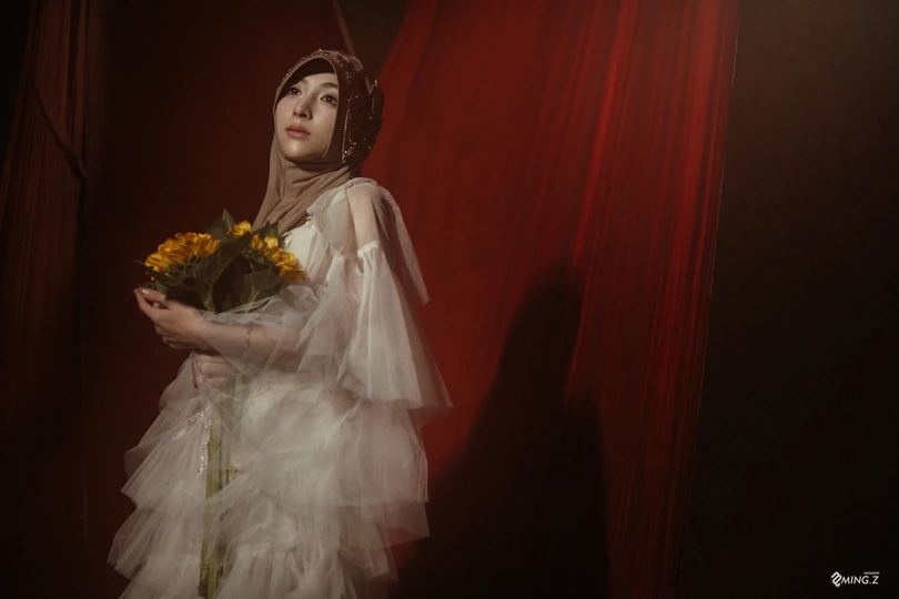 一位穿着白色连衣裙的年轻女子手持一束黄玫瑰 站在红色窗帘前。