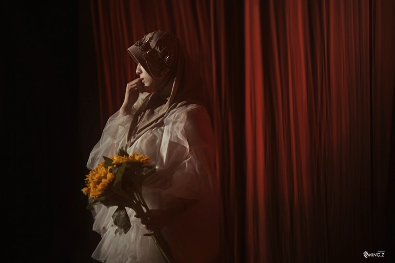一个戴白色面纱的女子手持一束黄色的花 站在红色 curtains 前。