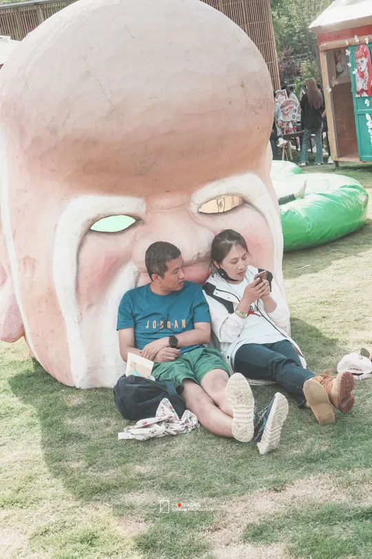 一个男子、一个女子和一个小女孩坐在草地上 旁边有一尊女性的脸部雕塑。