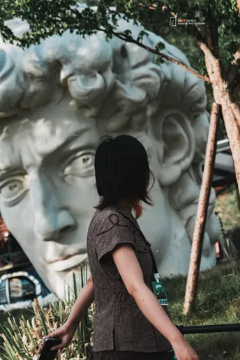 一个穿着连衣裙的小女孩站在一扇栅栏后面 带着一个箱子 正在注视着一个大型的人头雕塑。