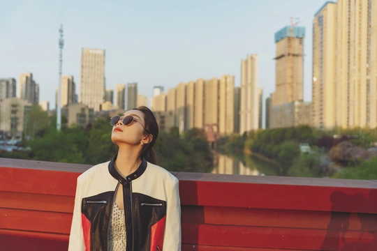 一个戴太阳镜的年轻女子坐在红色长凳上 背景是城市中的建筑。