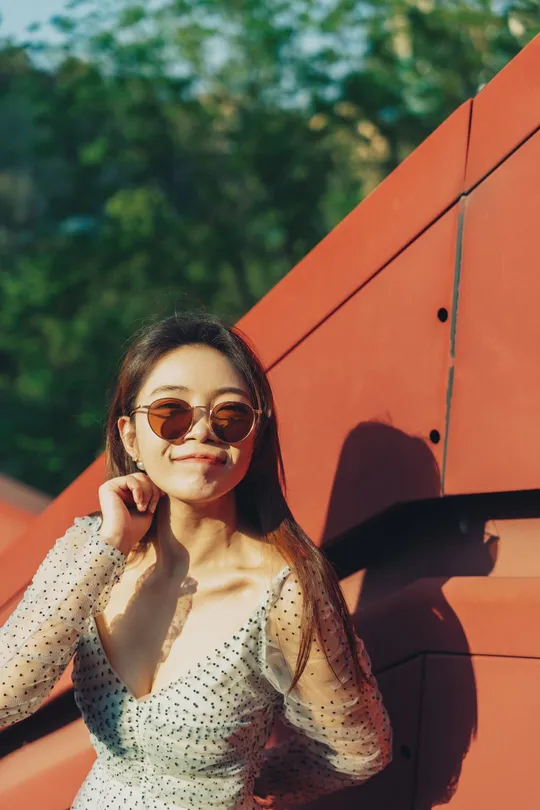 一位戴着太阳镜和连衣裙的年轻女子站在红色墙壁旁边