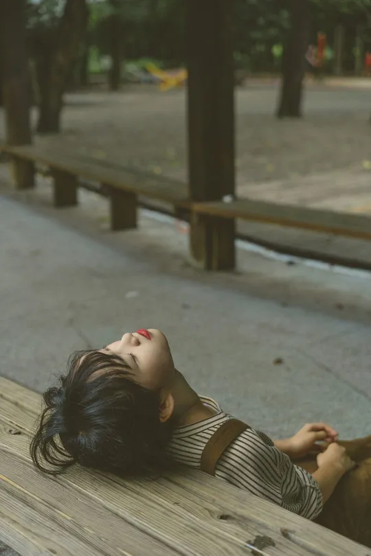 一位年轻女子在公园的木凳上躺着 把头靠在上面。