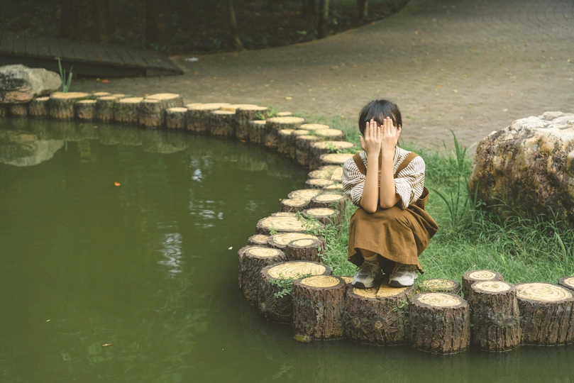 一个女孩坐在池塘边上 双手捂住脸 后面有一个人