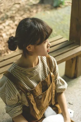 一个小女孩坐在木凳上 正在看一个小布绒玩具。