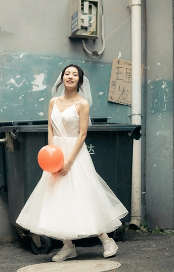 一位穿着白色连衣裙的年轻女子拿着一个橙色气球站在街上。