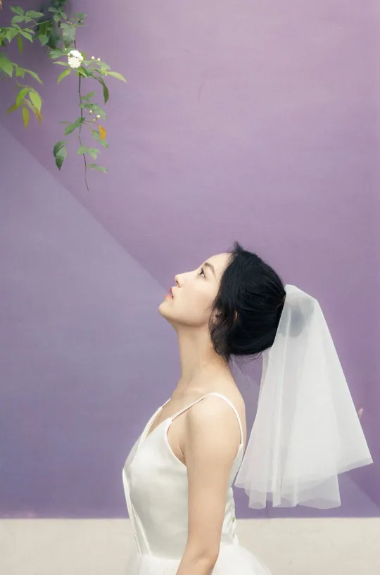 一位穿着白色长裙和面纱的年轻女子站在紫色的墙前