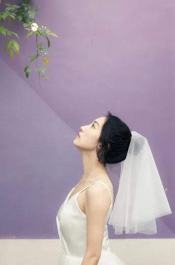 一位穿着白色长裙和面纱的年轻女子站在紫色的墙前