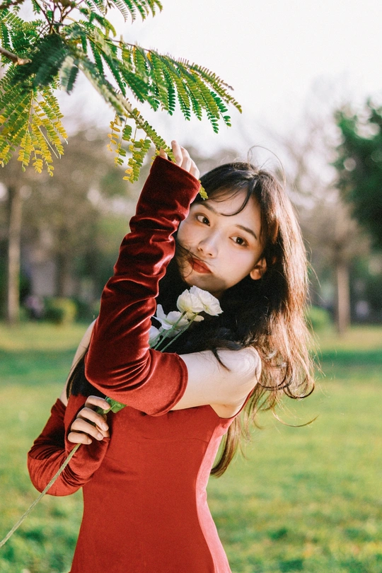 一位穿着红裙子的女士 戴着手套 手里拿着一朵花 站在一棵树旁边。