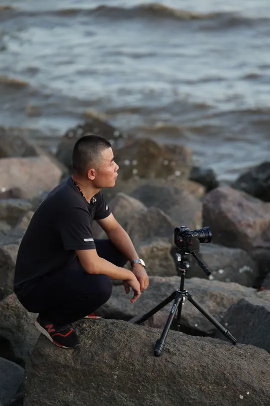 一个人坐在一些岩石上靠近海洋 他的相机放在三脚架上 正在给海滩拍照。