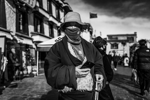 一张黑白照片 一位老人戴着面具 手拿拐杖在街上行走。