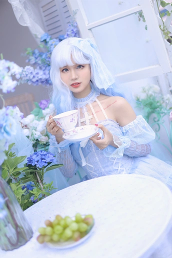 一个蓝头发的女人坐在一张白色的桌子旁 手里拿着一碗葡萄和一杯茶