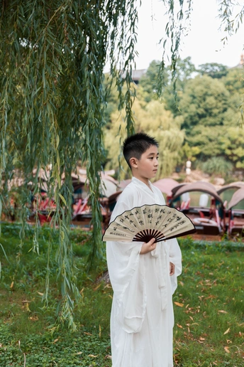 一个穿着白色长袍、打着雨伞的小男孩站在公园的柳树下