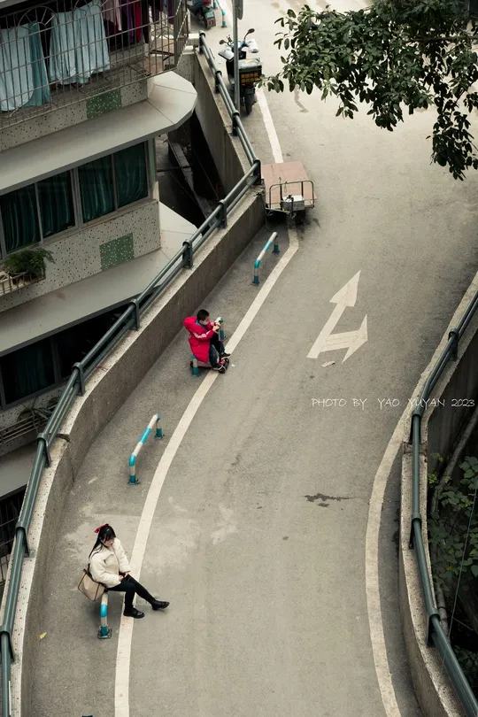 从上方视角看一条街道 一名男子坐在长凳上 一名女子骑着滑板车沿着道路中间行驶。