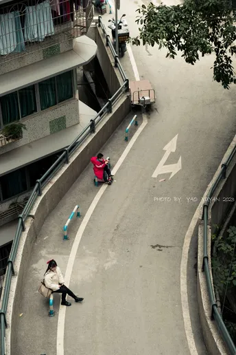 从上方视角看一条街道 一名男子坐在长凳上 一名女子骑着滑板车沿着道路中间行驶。