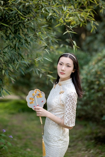 一位穿着传统白色裙子的年轻女子手持扇子 手握花 站在一棵树前。