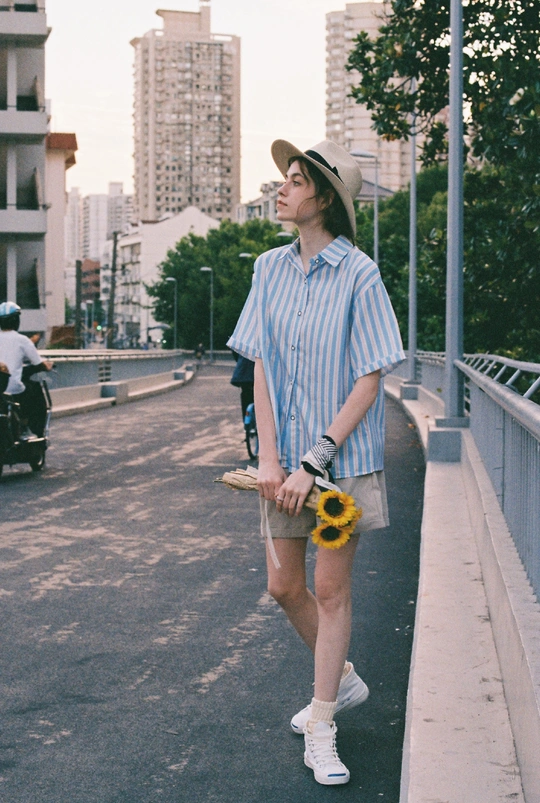 一个戴帽子的小伙子拿着向日葵和滑板走在街道人行道上。