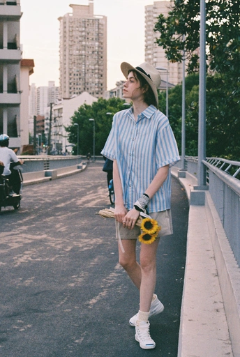 一个戴帽子的小伙子拿着向日葵和滑板走在街道人行道上。