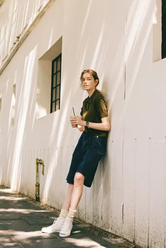 一位年轻女子站在人行道上 靠在一面白色墙壁旁 正在看手机。