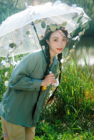 一位穿着绿色衣服的年轻女子在河边草地上举着一把透明的伞
