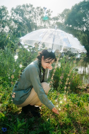 一位年轻女子手持透明雨伞坐在池塘边的田野上