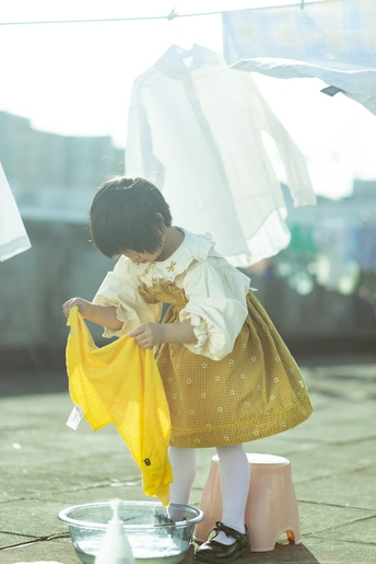 一个穿着黄色连衣裙的年轻女孩抱着一个小娃娃 她站在洗衣线上 地上放着一排要洗的衣服