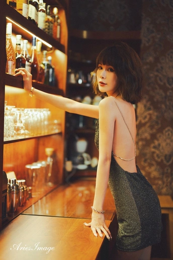 一个穿着连衣裙的年轻女子站在酒吧里 手里拿着瓶子和玻璃杯。