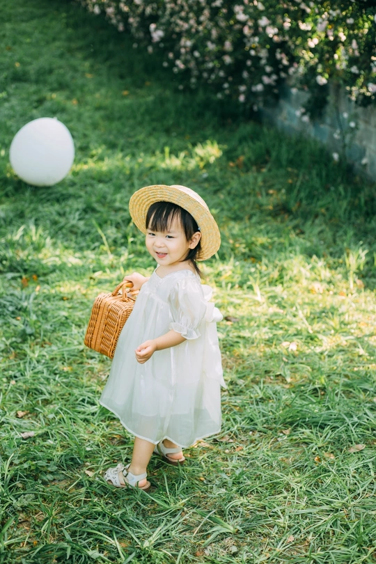 一个穿着白色连衣裙、戴着草帽的小女孩手里拿着一个装着球的篮子