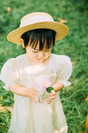 一个穿着白色连衣裙、戴着草帽的小女孩闻到了草丛中一朵粉红色的花的味道