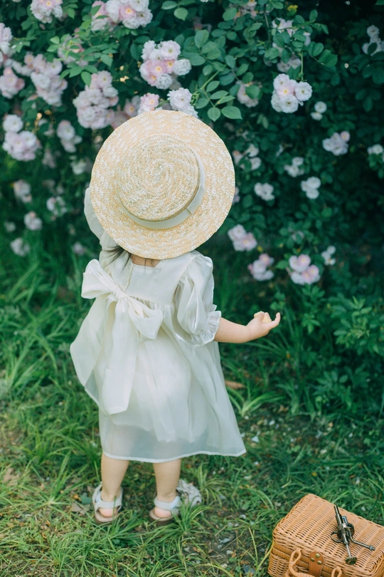 一个穿着连衣裙、戴着帽子的小女孩站在花园里一个手提箱旁边的草地上