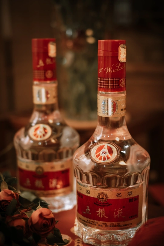 一瓶酒坐在桌子旁边 旁边有一杯玫瑰花。