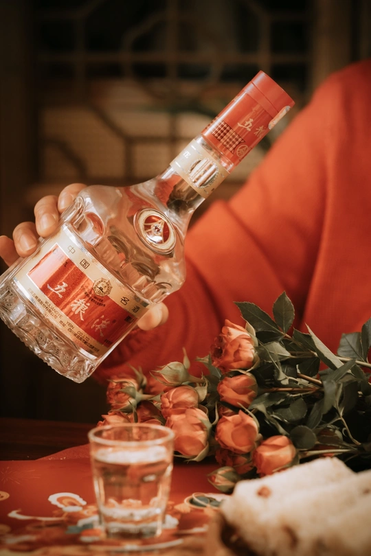 一位女性手持一瓶伏特加酒 旁边放着一杯含酒精的饮料 放在有花的桌子旁边。