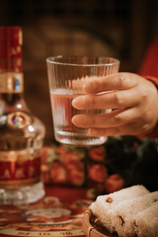 一个人手里拿着一杯酒 背景有一个盘子和一瓶酒精饮料。