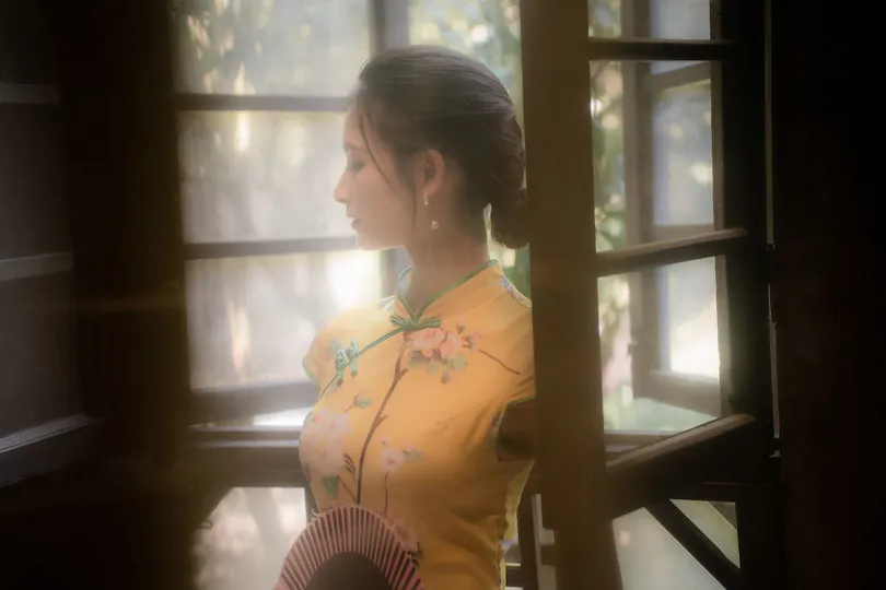 一位穿着黄色连衣裙的年轻女子拿着扇子 正凝视着打开的窗户。