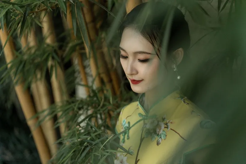 一位穿着黄色连衣裙的年轻女子站在竹林中 周围有绿色的植物和一把伞。