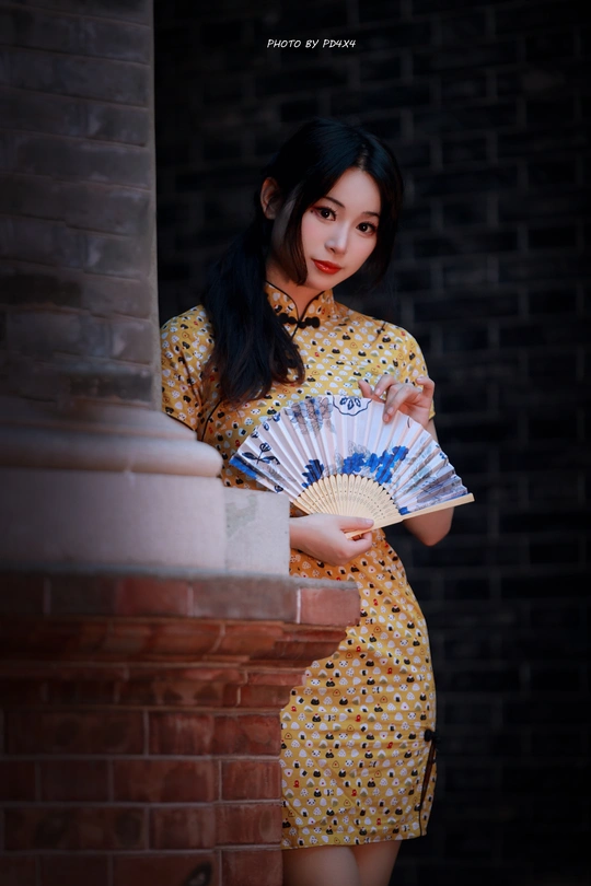 一个穿着黄色连衣裙的年轻女子手里拿着一把扇子 站在砖墙旁