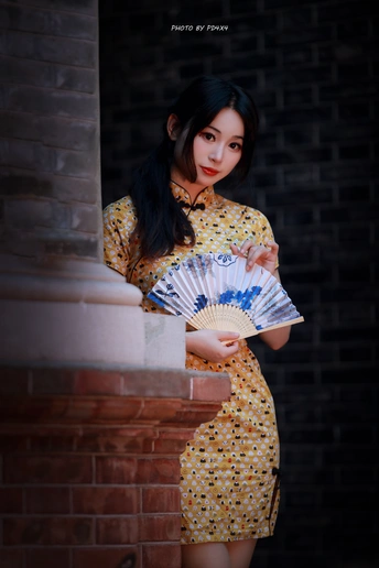 一个穿着黄色连衣裙的年轻女子手里拿着一把扇子 站在砖墙旁
