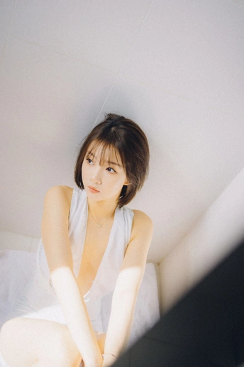 一个穿着白色连衣裙的年轻女子坐在床上 对着镜子看。