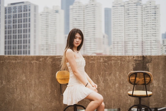 一个穿着白色连衣裙的年轻女子站在阳台上 可以俯瞰城市 阳台上有一堵墙 墙上有椅子。