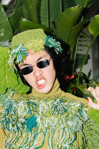 一位戴着太阳镜和绿色帽子的女人伸出舌头 在一片绿色的叶状植物前摆姿势。