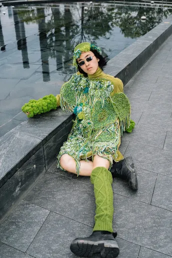 一位穿着绿色连衣裙和绿色靴子的女人坐在绿色毛绒玩具旁边地上。